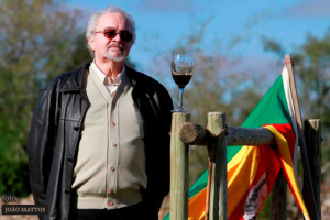 Jornalista Danilo Ucha ao lado de uma porteira com um taça de vinho e a bandeira do Rio Grande do Sul. Foto de João Mattos