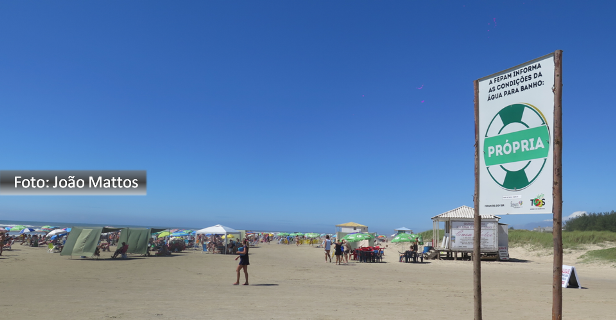 Foto de João Mattos mostra a praia de Atlântida com céu azul, pessoas na orla e placa de balneabilidade