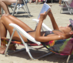 Mulher de biquíni lê um livro à beira-mar, deitada de costas em sua cadeira de praia
